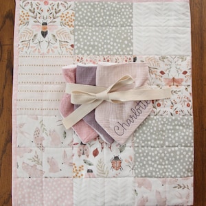 May include: A pink and gray patchwork baby quilt with a floral design and a set of four muslin burp cloths tied with a beige ribbon. The burp cloths are embroidered with the name "Charlotte".