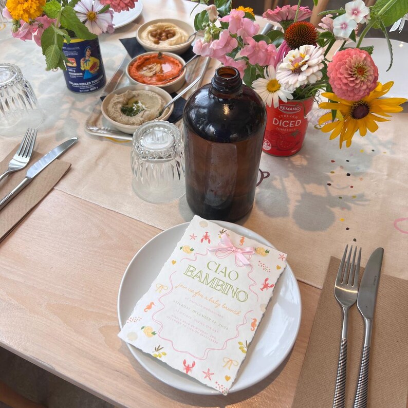 May include: A pink and white baby shower invitation with the text "Ciao Bambino" and a pink ribbon. The invitation is on a white plate with a floral arrangement in the background.