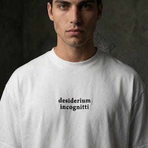 Pode incluir: Camiseta branca com as palavras "desiderium incognitti" impressas em preto. A camisa é feita de um material macio e tem gola redonda. A camisa é um item de roupa casual.