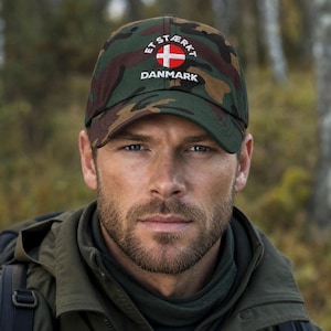 May include: A camouflage baseball cap featuring a circular Danish flag emblem in the centre. The text ET STÆRKT is embroidered in white above the flag, and the word DANMARK is embroidered in white below the flag on the front panel.