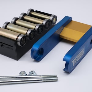 Può includere: Un assortimento di parti di macchine metalliche, tra cui rulli, staffe e bulloni. I rulli sono dorati e argentati, inseriti in un telaio nero con il testo "TR MAKER". Sono presenti anche staffe blu e bulloni argentati.