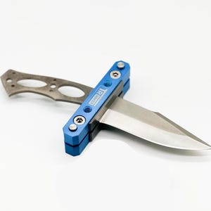 Peut inclure: Un couteau de poche bleu et argent avec une lame partiellement ouverte. Le couteau porte le logo TRIWAKER sur le côté.