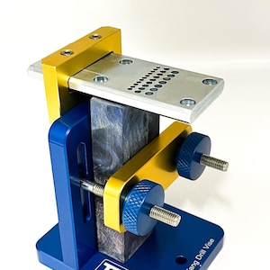 Peut inclure: Un étau de perçage bleu et doré avec une plaque supérieure argentée et des boutons bleus. L'étau maintient un bloc de bois avec un motif bleu et noir. Le texte "TR MAKER" est visible sur la base.