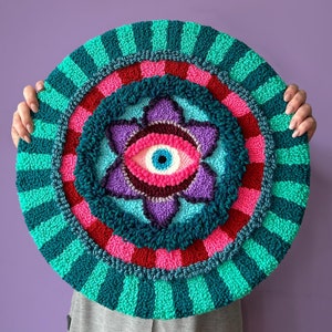 May include: A colorful, round wall hanging with a large, stylized eye in the center. The eye is surrounded by a flower-like design with pink, purple, and blue petals. The background is a series of concentric circles in shades of green, pink, and red.