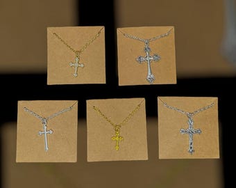 May include: Five cross necklaces displayed on small brown cards. The crosses are in gold and silver tones, with varying designs. Some have ornate detailing, while others are simple. The necklaces are on delicate chains.