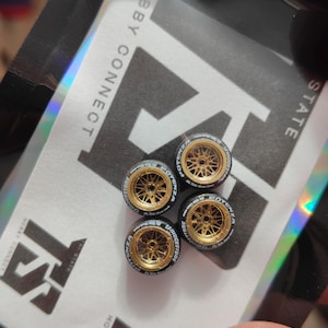 Może przedstawiać: Cztery miniaturowe złote felgi aluminiowe z czarnymi oponami. Opony mają białe napisy z napisami "POTENZA" i "BRIDGESTONE". Koła znajdują się na holograficznym opakowaniu z napisem "HOBBY CONNECT" i "HI-STATE".