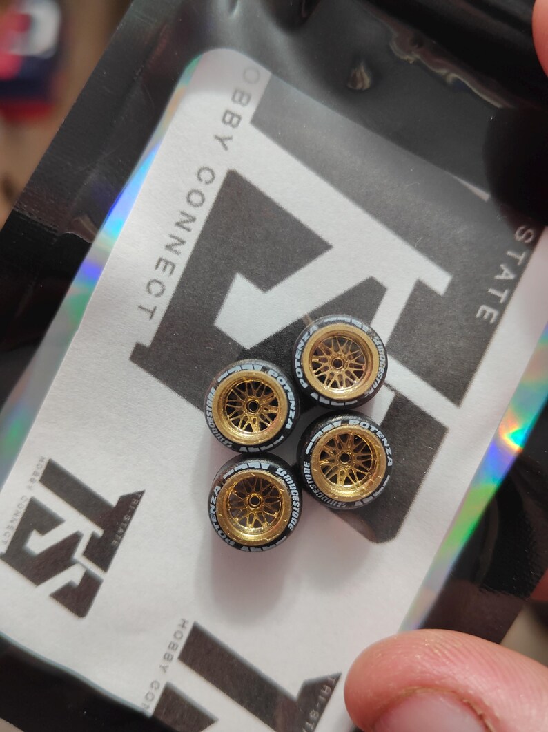 Może przedstawiać: Cztery miniaturowe złote felgi aluminiowe z czarnymi oponami. Opony mają białe napisy z napisami "POTENZA" i "BRIDGESTONE". Koła znajdują się na holograficznym opakowaniu z napisem "HOBBY CONNECT" i "HI-STATE".
