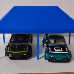 May include: A blue miniature car shelter with two toy cars parked underneath. One car is black with a green stripe, and the other is teal with the number 73 on the hood. The shelter is a bright blue color.