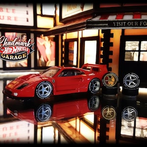 May include: A red toy car with silver rims and a rear spoiler is parked in front of a miniature garage. The garage has a sign that says "Visit Our Fountain". The logo "Chadmark Hot Wheels Garage" is visible.