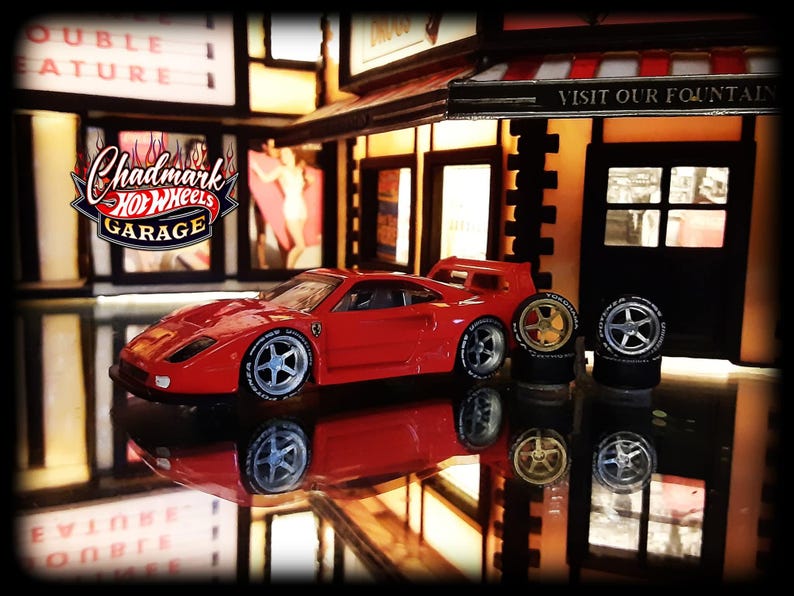 May include: A red toy car with silver rims and a rear spoiler is parked in front of a miniature garage. The garage has a sign that says "Visit Our Fountain". The logo "Chadmark Hot Wheels Garage" is visible.