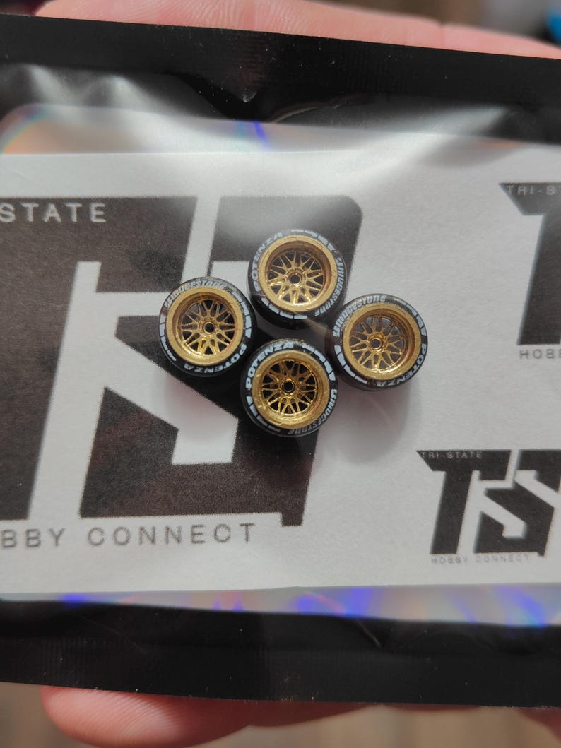 Może przedstawiać: Cztery miniaturowe złote felgi aluminiowe z czarnymi oponami. Koła mają wieloramienną konstrukcję i są oznaczone słowami "POTENZA" i "BRIDGESTONE". Koła te są przeznaczone do modelu samochodu lub podobnego przedmiotu hobbystycznego.
