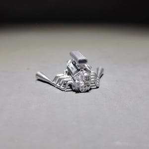 Puede incluir: Un pequeño modelo plateado de un motor V8. El motor tiene componentes detallados, incluyendo tubos de escape, un carburador y bujías. El modelo está sobre un fondo gris.