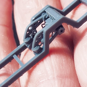 Puede incluir: Primer plano de un componente de chasis de modelo de plástico gris. El componente presenta detalles intrincados, incluyendo brazos curvos, aberturas circulares y una serie de pequeñas piezas cilíndricas. El diseño sugiere un tema mecánico.