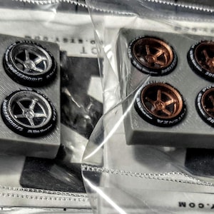 Puede incluir: Dos juegos de ruedas de coche en miniatura. Uno tiene llantas plateadas y neumáticos negros con letras blancas. El otro tiene llantas de bronce y neumáticos negros. Cada juego tiene cuatro ruedas.