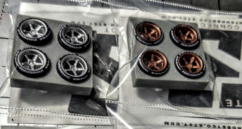 May include: Two sets of miniature car wheels. One set has silver rims and black tyres with white lettering. The other set has bronze rims and black tyres with white lettering. Each set has four wheels.