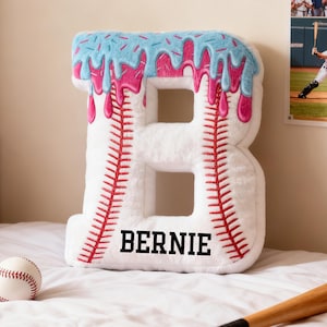 May include: A white, plush letter "B" cushion, designed like a baseball with red stitching. It features blue sprinkles and pink icing at the top. The name "BERNIE" is printed in black. A baseball and a wooden bat are also visible.