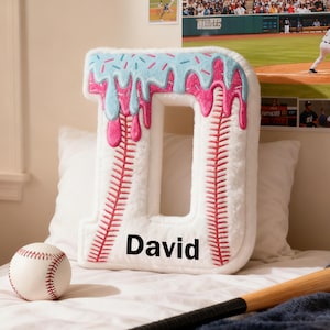 Op de afbeelding: Een zacht, wit kussen in de vorm van de letter "D" met roze en blauwe glazuurdetails, die op een donut lijken. Het kussen heeft rode stiksels, een honkbalontwerp en de naam "David" erop gedrukt. Een honkbal en een knuppel zijn ook zichtbaar.