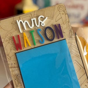 Pu&ograve; includere: Un blocco note in legno con il nome "Mrs. Watson" in lettere colorate. Include un blocco di note adesive blu e una matita. Lo sfondo &egrave; decorato con illustrazioni a tema scolastico.