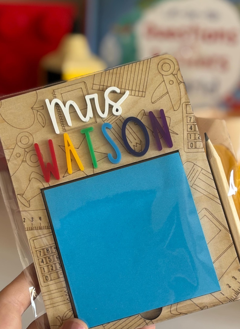 Pu&ograve; includere: Un blocco note in legno con il nome "Mrs. Watson" in lettere colorate. Include un blocco di note adesive blu e una matita. Lo sfondo &egrave; decorato con illustrazioni a tema scolastico.