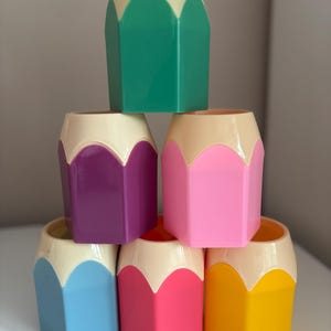 May include: A stack of six hexagonal pencil-shaped pen holders made of plastic. The top section of each holder is cream-colored, while the bottom sections are colored green, purple, pink, light blue, hot pink, and yellow respectively.