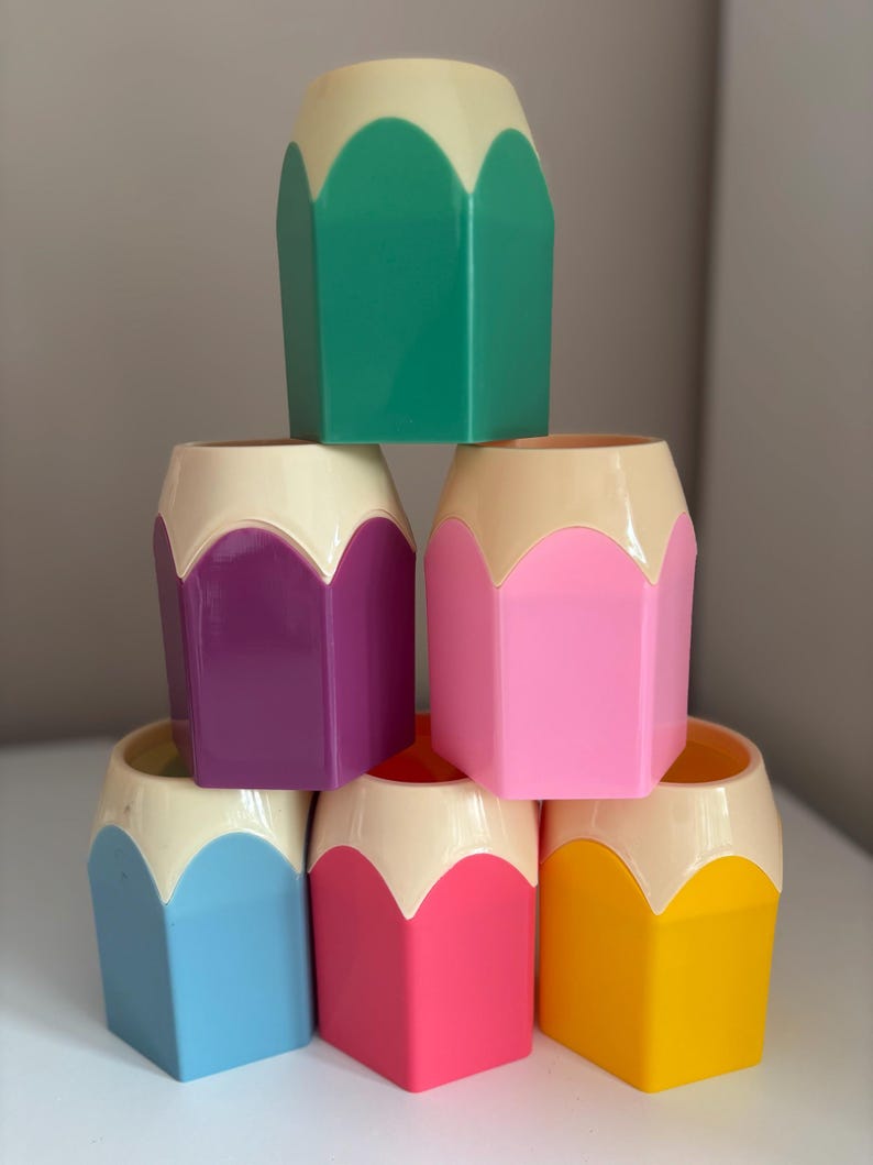 May include: A stack of six hexagonal pencil-shaped pen holders made of plastic. The top section of each holder is cream-colored, while the bottom sections are colored green, purple, pink, light blue, hot pink, and yellow respectively.
