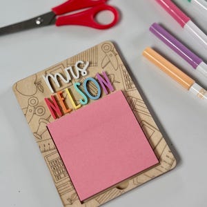 Pu&ograve; includere: Un portablocco in legno con il nome "Mrs. Nelson" in lettere colorate. Il blocco note ha un blocco note adesivo rosa ed &egrave; decorato con illustrazioni a tema scolastico. Sono visibili anche forbici rosse e pennarelli colorati.