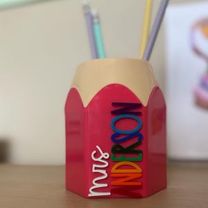 May include: A pink pencil-shaped desk organizer with a beige top section. The side features the white text "mrs" and the name "ANDERSON" in stacked, multi-colored letters including blue, green, yellow, orange, and red. Several pencils are inside.