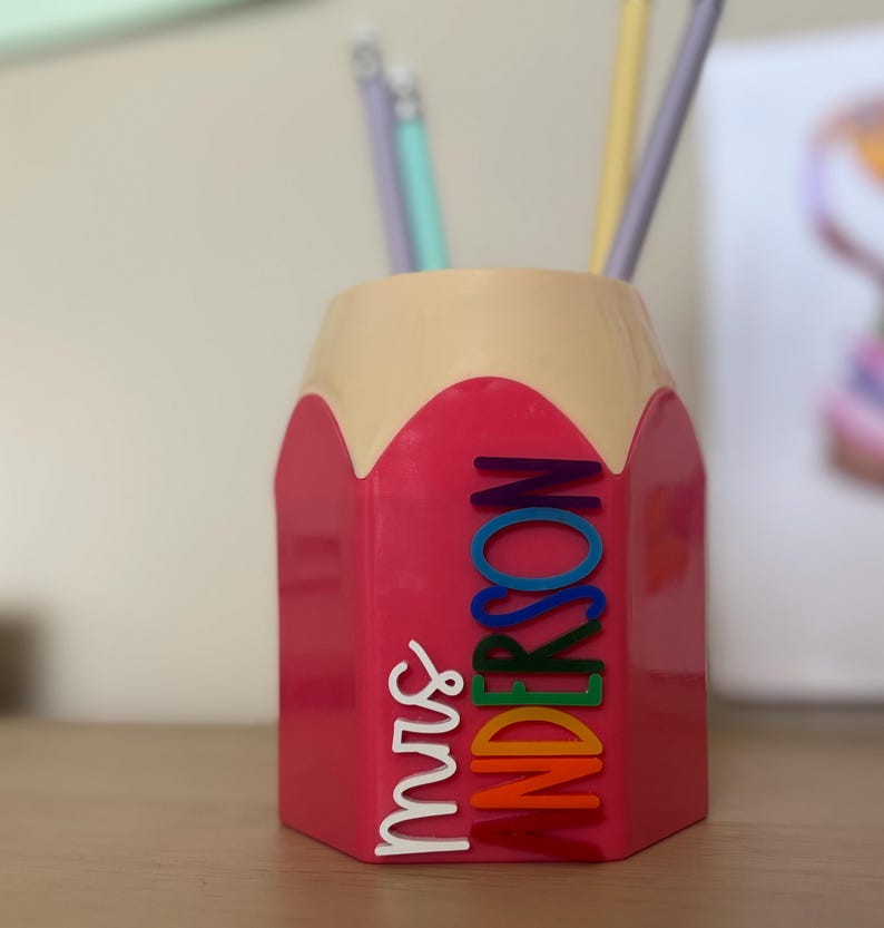 May include: A pink pencil-shaped desk organizer with a beige top section. The side features the white text "mrs" and the name "ANDERSON" in stacked, multi-colored letters including blue, green, yellow, orange, and red. Several pencils are inside.