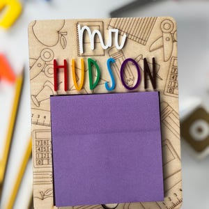 Pu&ograve; includere: Organizzatore da scrivania per insegnanti in legno con il nome "HUDSON" in lettere colorate e "mr" in bianco. L'organizzatore ha un blocco note viola ed &egrave; decorato con illustrazioni a tema scolastico, tra cui matite, forbici e mele.