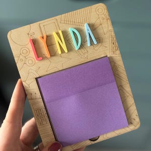 Pu&ograve; includere: Una lavagna in legno con il nome "LYNDA" in lettere 3D colorate. La lavagna ha un blocco note adesivo viola ed &egrave; decorata con disegni incisi di articoli per la scuola. La lavagna &egrave; tenuta da una persona.