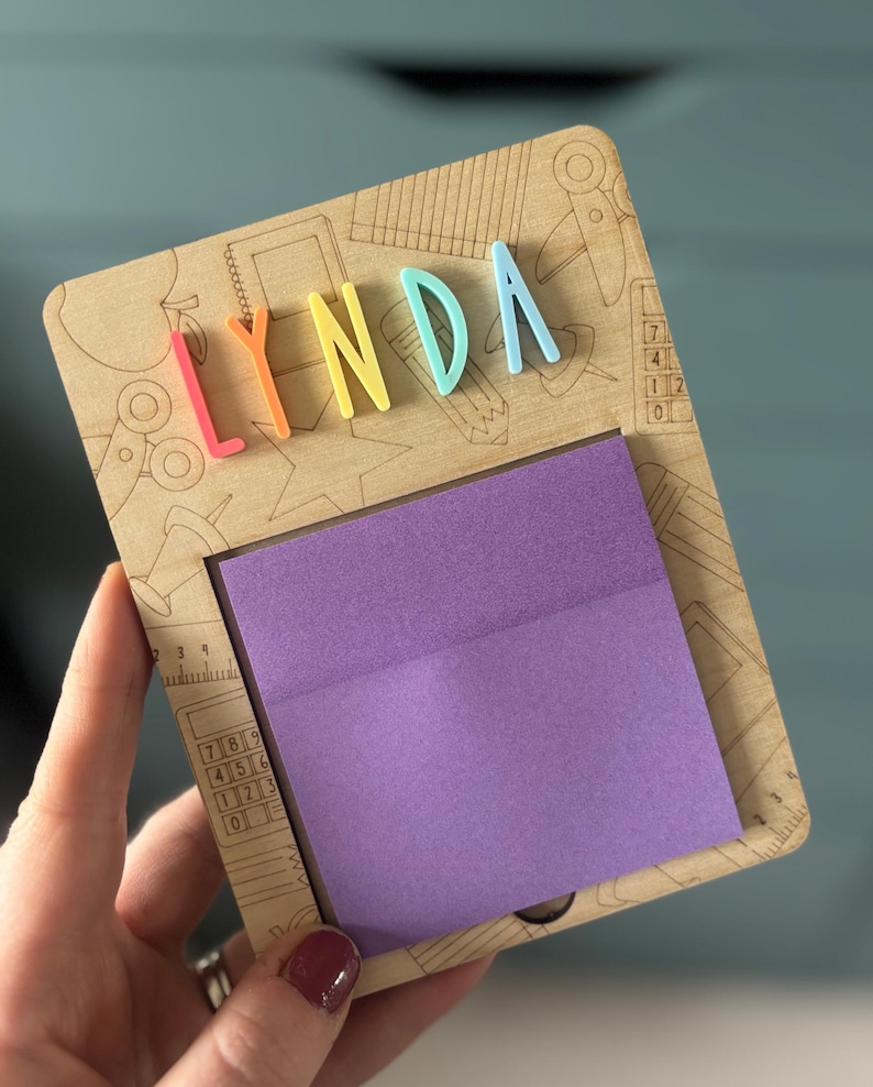 Pu&ograve; includere: Una lavagna in legno con il nome "LYNDA" in lettere 3D colorate. La lavagna ha un blocco note adesivo viola ed &egrave; decorata con disegni incisi di articoli per la scuola. La lavagna &egrave; tenuta da una persona.