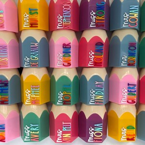 May include: A collection of pencil-shaped desk organizers in various colors including pink, yellow, blue, green, and purple. Each organizer features a beige tip and a personalized name tag with titles like Mrs, Miss, or Ms in colorful lettering.