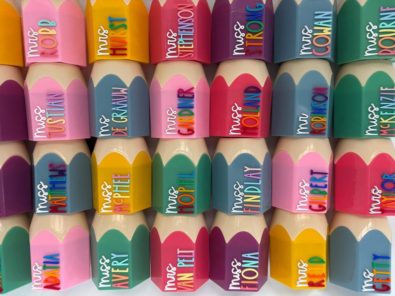 May include: A collection of pencil-shaped desk organizers in various colors including pink, yellow, blue, green, and purple. Each organizer features a beige tip and a personalized name tag with titles like Mrs, Miss, or Ms in colorful lettering.
