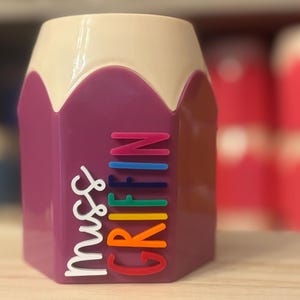 May include: A purple pencil-shaped desk organizer with a cream-colored top. The side features the text "Miss GRIFFIN" in raised, multi-colored lettering, with "Miss" in white and "GRIFFIN" in stacked, vibrant rainbow-colored letters.