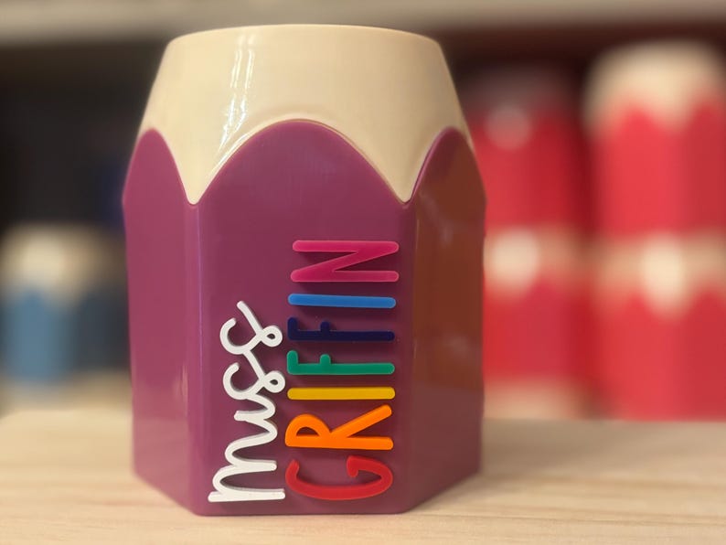 May include: A purple pencil-shaped desk organizer with a cream-colored top. The side features the text "Miss GRIFFIN" in raised, multi-colored lettering, with "Miss" in white and "GRIFFIN" in stacked, vibrant rainbow-colored letters.