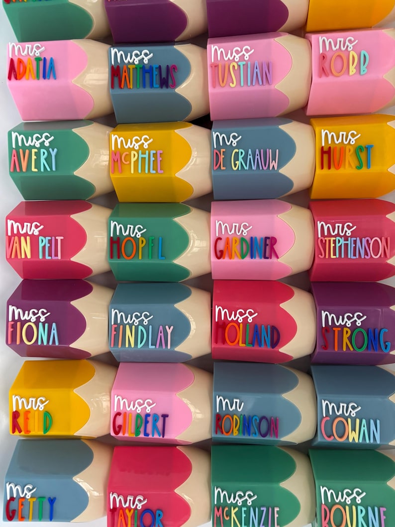 May include: A collection of pencil-shaped containers in various colors, each featuring a raised white title such as Mrs, Miss, or Ms, followed by a name in colorful, individual letters. The containers have a beige tip and a hexagonal body.