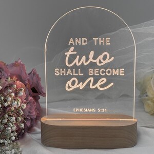 May include: A clear acrylic sign with a wooden base, featuring the text "And the two shall become one" with the scripture reference "Ephesians 5:31".