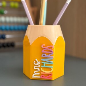 May include: A yellow pencil-shaped desk organizer with a light peach top section. The side features the name "mrs RICHARDS" in raised, multi-colored lettering. Three pencils are placed inside the container, which has a hexagonal base.