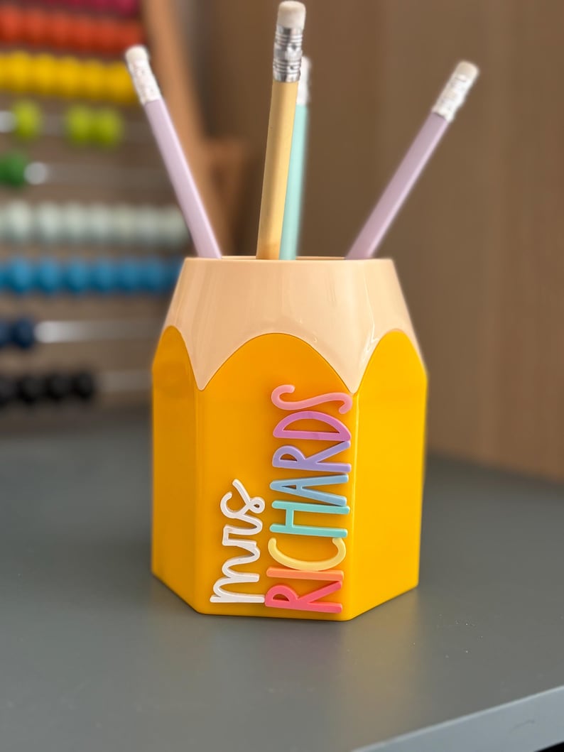 May include: A yellow pencil-shaped desk organizer with a light peach top section. The side features the name "mrs RICHARDS" in raised, multi-colored lettering. Three pencils are placed inside the container, which has a hexagonal base.