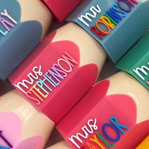May include: A collection of pencil-shaped containers in various colors including pink, blue, and yellow. Each container features a white script title such as mrs STEPHENSON or mr ROBINSON, followed by the surname in raised, multi-colored lettering.