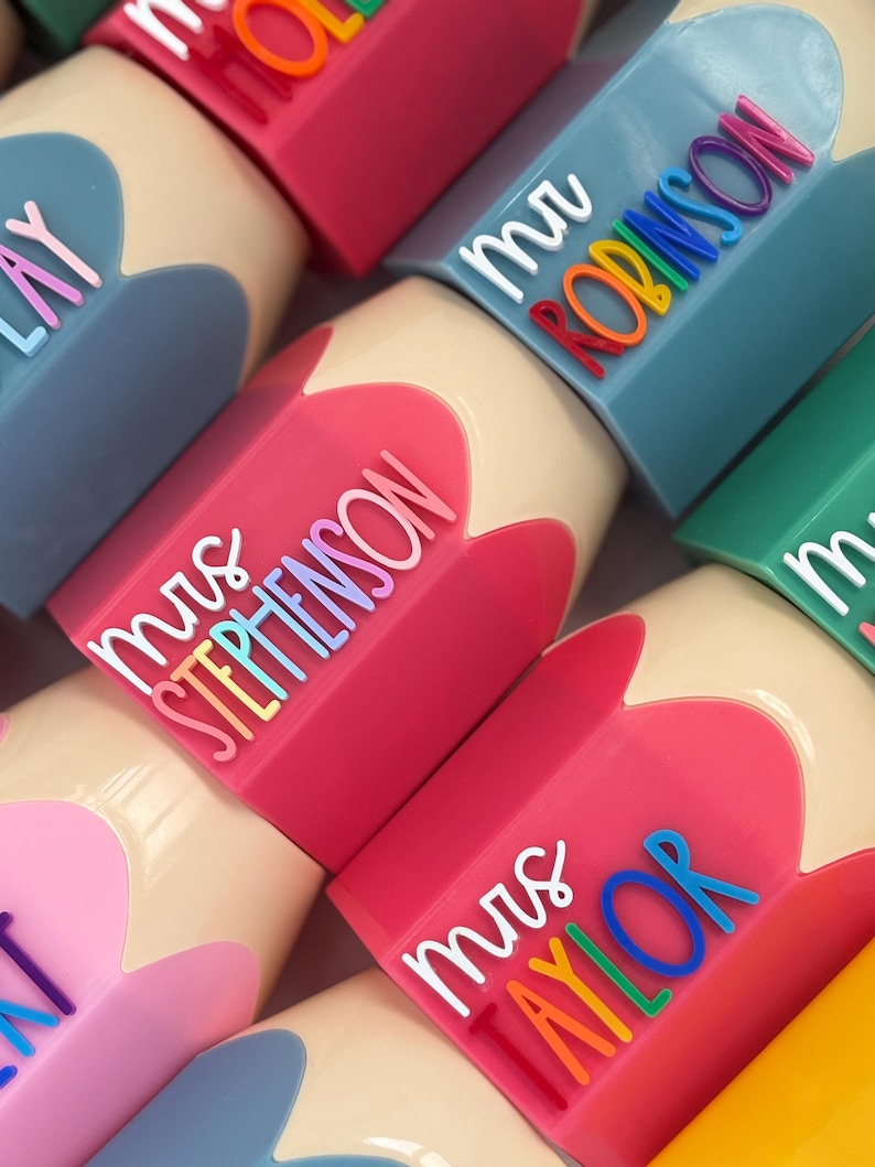 May include: A collection of pencil-shaped containers in various colors including pink, blue, and yellow. Each container features a white script title such as mrs STEPHENSON or mr ROBINSON, followed by the surname in raised, multi-colored lettering.