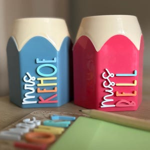 May include: Two pencil-shaped desk organizers with cream-colored tops. The left organizer is blue with the white, yellow, and coral text "mrs KEHOE". The right organizer is pink with the white, yellow, and coral text "miss BELL".