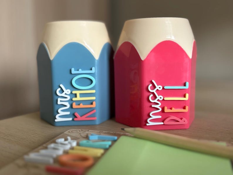 May include: Two pencil-shaped desk organizers with cream-colored tops. The left organizer is blue with the white, yellow, and coral text "mrs KEHOE". The right organizer is pink with the white, yellow, and coral text "miss BELL".