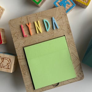 Pu&ograve; includere: Portablocco in legno con il nome "LYNDA" in lettere colorate. Il supporto ha un blocco note adesivo verde ed &egrave; circondato da blocchi di lettere in legno. Il supporto &egrave; di colore marrone chiaro con disegni incisi.