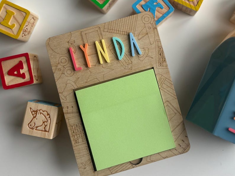 Pu&ograve; includere: Portablocco in legno con il nome "LYNDA" in lettere colorate. Il supporto ha un blocco note adesivo verde ed &egrave; circondato da blocchi di lettere in legno. Il supporto &egrave; di colore marrone chiaro con disegni incisi.