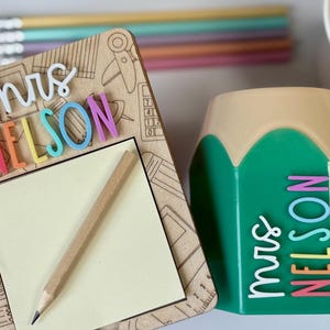 May include: A wooden desk organiser with the name "Mrs. Nelson" in colourful letters, a pencil, and a notepad. A green pencil-shaped container with the name "Mrs. Nelson" is also visible. Pencils are in the background. The items are likely for a teacher.
