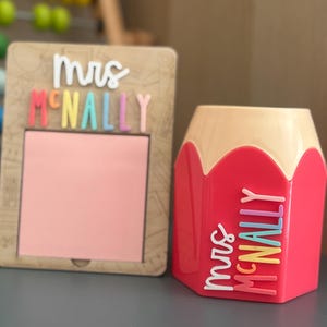 May include: A wooden square memo holder with a pink square insert and the white and multi-colored text "Mrs McNally" above it, paired with a pink and beige pencil-shaped pen holder featuring the white and multi-colored text "Mrs McNally".