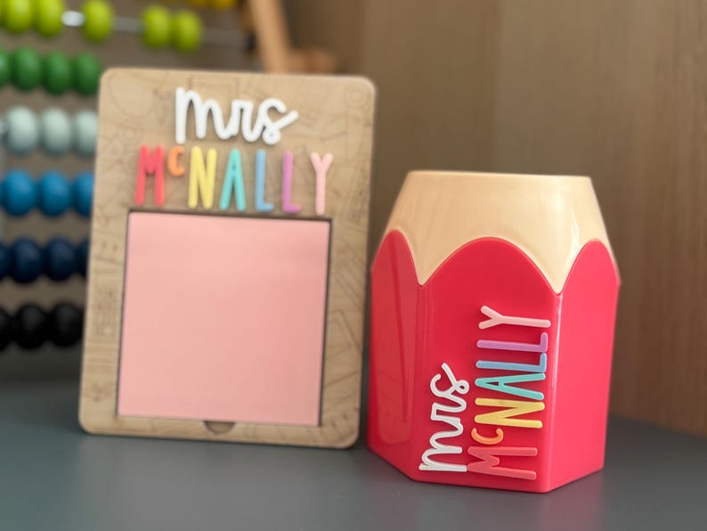 May include: A wooden square memo holder with a pink square insert and the white and multi-colored text "Mrs McNally" above it, paired with a pink and beige pencil-shaped pen holder featuring the white and multi-colored text "Mrs McNally".