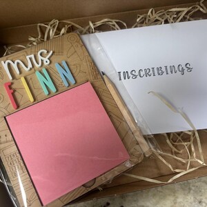 Pu&ograve; includere: Un set regalo a tema insegnante in una scatola di cartone. Include una cornice in legno con il nome "Mrs. Finn" in lettere colorate, un blocco note rosa, una matita e una busta bianca con la scritta "Inscribings".