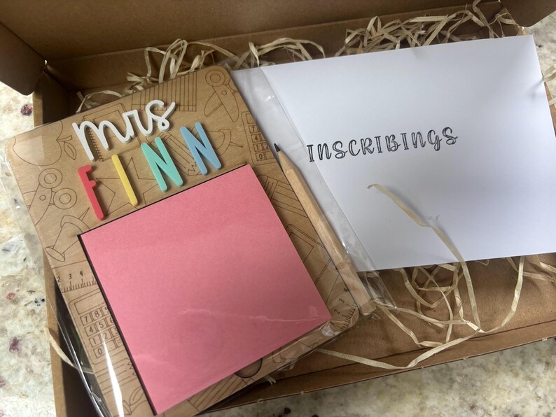 Pu&ograve; includere: Un set regalo a tema insegnante in una scatola di cartone. Include una cornice in legno con il nome "Mrs. Finn" in lettere colorate, un blocco note rosa, una matita e una busta bianca con la scritta "Inscribings".